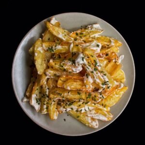 Top view of golden potato wedges drizzled with creamy sauce and herbs on a black background.